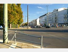 Straßenkreuzung in einer Stadt mit mehrspurigen Fahrbahnen, parkenden und fahrenden Autos. Auf der linken Seite stehen Bäume mit herbstlichem Laub, rechts sind mehrstöckige Wohn- und Geschäftshäuser mit Balkonen und Geschäften im Erdgeschoss sichtbar.