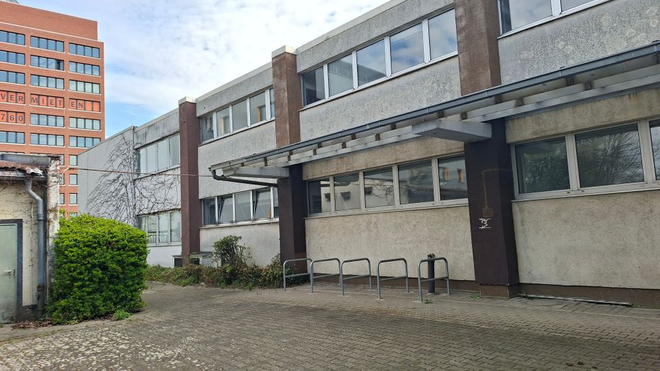 Innenhof vor einem flachen, zweigeschossigen Büro- oder Industriegebäude mit grauer Putzfassade, langen Fensterbändern und einem auskragenden Vordach über dem Eingang. Vor dem Gebäude stehen Metall-Fahrradbügel auf einer gepflasterten Fläche, daneben Büsche und Kletterpflanzen; im Hintergrund ragt ein hoher Backsteinbau auf.