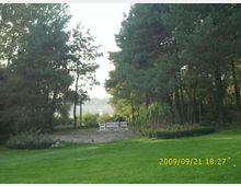 Grüne Gartenlandschaft mit gepflegtem Rasen, hohen Nadelbäumen im Hintergrund und einem weißen Gartenmöbel-Set aus zwei Bänken und einem Tisch in der Mitte. Im Hintergrund ist ein See hinter den Bäumen erkennbar.