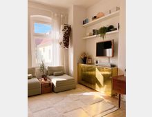 Helles Wohnzimmer mit großen Fenstern, weißen Vorhängen und zwei beigefarbenen Sesseln neben einem Holztisch. Eine goldene Kommode, zwei Wandregale mit Büchern und Dekorationen sowie ein an der Wand montierter Fernseher ergänzen die Einrichtung.