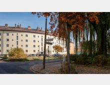 Kreuzung mit Straßenschildern für „Holzkircher Straße“ und „Gablerstraße“, umgeben von herbstlich gefärbten Bäumen. Im Hintergrund steht ein mehrstöckiges Wohngebäude mit heller Fassade und zahlreichen Fenstern, daneben parkende Autos.