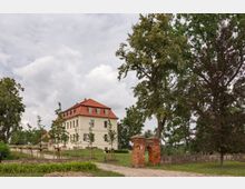 Eine historische Villa mit cremefarbener Fassade und rotem Mansarddach steht auf einem gepflegten Grundstück mit gepflastertem Weg und jungen Bäumen. Im Vordergrund ist ein kleines, aus roten Ziegeln gemauertes Torbogenfragment zu sehen, umgeben von Rasen und hohen Bäumen.