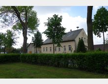 Gelbes Gebäude mit Satteldach und mehreren weißen Fenstern, umgeben von Bäumen und einer geschnittenen Hecke im Vordergrund. Die Szene befindet sich in einer grünen, ländlichen Umgebung unter bewölktem Himmel.