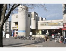 Ein moderner Platz mit einem weißen, geometrischen Gebäude im Hintergrund, das für Geschäfte und Studios genutzt wird. Vor dem Gebäude befinden sich ein Fahrradständer, eine Straßenbeschilderung mit der Aufschrift „Richard-Wagner-Platz“, sowie ein Gehweg mit Passanten und Sitzbereichen eines Straßencafés.