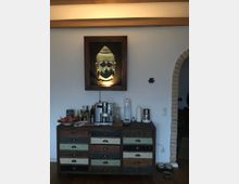 Rustikale Wohnzimmerecke mit einer Kommode aus Holz und bunten Schubladen. Auf der Kommode stehen eine Kaffeemaschine, eine Karaffe, ein Wassersprudler und verschiedene Flaschen; darüber hängt ein Buddha-Bild mit Beleuchtung an einer weißen Wand.