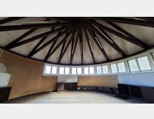 Innenraum eines runden Pavillons mit kuppelförmiger Decke, deren dunkle Balken strahlenförmig zur Mitte verlaufen. Entlang der gebogenen Wand befindet sich ein umlaufendes Band aus kleinen Fenstern über einer niedrigen Sitzbank und Lüftungsgittern.