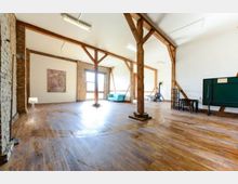 Geräumiger Innenraum mit Holzboden und freiliegenden Holzbalken, teils mit Ziegelwänden und weißen Wänden gestaltet. Der Raum enthält ein großes Fenster mit Tageslichteinfall, ein blaues Sofa, einen Wandofen und eine grüne Tafel.