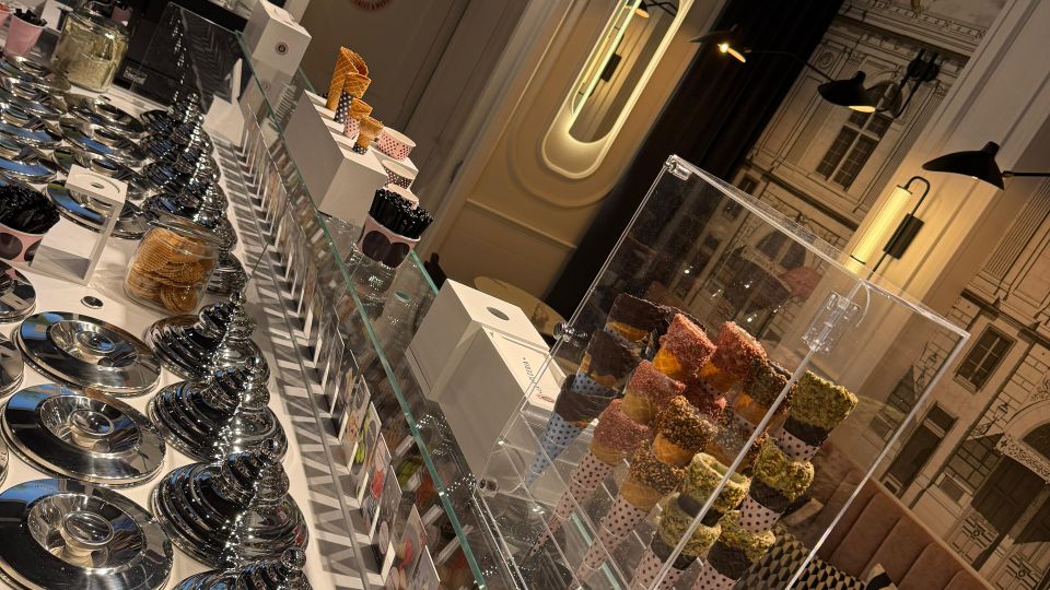 Eisdiele mit moderner Einrichtung: Im Vordergrund eine Glasvitrine mit silbernen Behältern für Eiscreme und dekorativen Eistüten in verschiedenen Farben. Im Hintergrund eine stilisierte Wandgestaltung, gepolsterte Sitzbänke mit Kissen und ein beleuchteter Werbebildschirm.