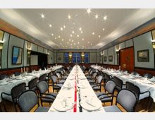 Eleganter Speisesaal mit zwei langen, weiß gedeckten Tischen, die von gepolsterten Stühlen umgeben sind. An den Wänden hängen maritime Gemälde, und die Decke ist mit zahlreichen deckenleuchtenden Spots ausgestattet.