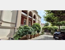 Innenhof mit Kopfsteinpflaster vor einem klassischen Gebäude mit hohen Säulen und Rundbogen-Arkaden. Große Pflanzkübel mit dichtem Grün säumen den Weg, der unter einer bewachsenen Pergola zu einem Tor und einem Nebengebäude führt.