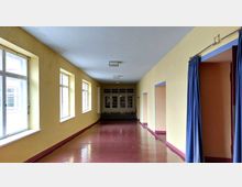 Ein heller Flur mit gelb gestrichenen Wänden, großen Fenstern auf der linken Seite und mehreren offenen Durchgängen rechts, teils mit blauen Vorhängen. Der Boden ist glänzend rot, und am Ende befindet sich eine geschlossene Tür mit Glasfenstern und Holzrahmen.