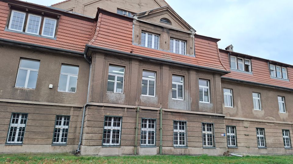 Alt-Text: Außenansicht eines mehrstöckigen, historischen Gebäudes mit brauner Fassade, weißen Fenstern und roten Ziegeldächern. Das Erdgeschoss zeigt große Gitterfenster, während die oberen Stockwerke kleinere Fenster und ein Giebeldach haben.
