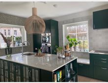 Moderne Küche mit dunkelgrünen Schränken und einer zentralen Kücheninsel mit weißer Marmor-Arbeitsfläche. Zwei große Fenster lassen Tageslicht herein, dekoriert mit Pflanzen und Küchenaccessoires; eine auffällige hölzerne Hängeleuchte hängt über der Insel.