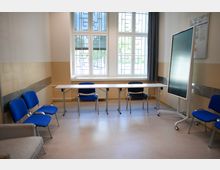 Ein kleiner, schlichter Besprechungsraum mit beigefarbenen Wänden, mehreren blauen Stühlen, einem rechteckigen Tisch und einem mobilen Bildschirm. Im Hintergrund befinden sich große Fenster mit Metallgittern, die Tageslicht hereinlassen.