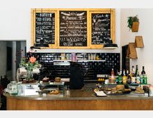 Eine gemütliche Café-Theke mit Holzverkleidung, schwarzer Fliesenrückwand und großen Kreidetafeln, die das Angebot an Getränken, Speisen und Snacks präsentieren. Auf der Theke stehen Kuchen, Croissants, Getränke und Dekorationen, darunter eine Blumenvasen.