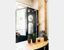 Heller Innenraum eines Cafés oder Arbeitsplatzes mit einem großen Holztisch, auf dem eine Ananas, Topfpflanzen und eine Schale stehen. Im Hintergrund sind eine schwarze Eingangstür mit Glaseinsätzen, Ziegelwände und gestapelte Stühle zu sehen.