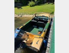 Kleines Motorboot mit Windschutzscheibe und Steuerstand, am Holzsteg in einem schmalen Uferbereich festgemacht. Im Hintergrund liegt eine gepflegte Rasenfläche mit Sträuchern und Gartenmöbeln am Ufer.
