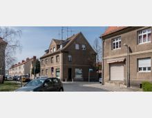 Straßenszene in einem Wohngebiet mit zweigeschossigen, sandfarbenen Gebäuden und Satteldächern. Im Vordergrund parken Autos, während Straßenlaternen und Schilder die Umgebung prägen; ein blauer Himmel und Bäume vervollständigen die Kulisse.