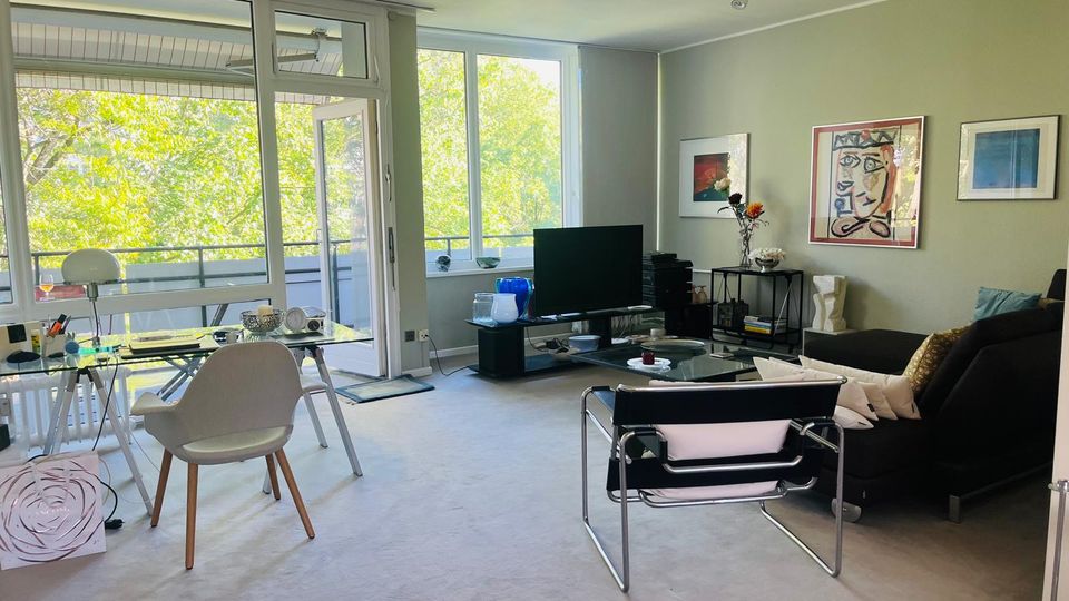 Helles, modern eingerichtetes Wohn- und Esszimmer mit großen Fensterfronten und offener Balkontür zum Balkon mit Blick auf grüne Bäume. Im Raum stehen ein Glastisch mit Stühlen, eine Sitzecke mit Sofa und Sessel sowie ein Fernseher; an der Wand hängen gerahmte Kunstwerke.