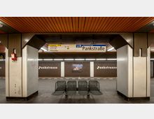 U-Bahn-Station mit der Bezeichnung „Pankstraße“ der Linie U8 in Berlin. Der Bahnsteig ist mit einer braunen Wandverkleidung, weißen Pfeilerschildern, Sitzbänken und einem Deckenpanel aus Holzoptik gestaltet; ein Hinweisschild oben zeigt Ausgänge und die Fahrtrichtung an.