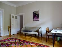 Wohnraum mit Holzboden und pastellfarbenen Wänden. Im Vordergrund liegt ein bunter Teppich mit geometrischem Muster, im Hintergrund stehen ein Sofa, ein Holzstuhl und ein Schreibtisch; an der Wand hängt ein gerahmtes Bild.