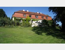 Ein zweistöckiges Landhaus mit weißer Fassade, roten Dachziegeln und mehreren Dachfenstern, umgeben von grünen Pflanzen und einer großen Rasenfläche im Vordergrund. Kletterpflanzen schmücken Teile der Fassade, und die Umgebung ist von Bäumen eingefasst.