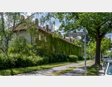 Straßenszene mit einem langgestreckten Wohnhaus, dessen Fassade mit dichtem Efeu bewachsen ist. Im Vordergrund befindet sich ein Gehweg mit Bäumen und einem Straßenschild mit der Aufschrift "Schlettstadter Straße", daneben parkende Autos.