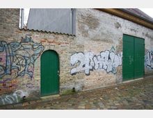Außenansicht einer alten Backsteinmauer mit grünen Türen, bedeckt von Graffiti. Der Boden davor besteht aus unregelmäßig gepflastertem Stein.
