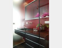 Modernes Badezimmer mit einer Kombination aus roten Wänden und dunklen Holzschränken. Auf dem Waschtisch steht ein Blumentopf mit einer lilafarbenen Orchidee, daneben zwei Trinkgläser; die Armatur ist minimalistisch gestaltet.