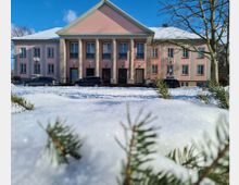 Ein rosafarbenes Gebäude mit klassizistischer Fassade und Säuleneingang, umgeben von schneebedecktem Boden und Tannenästen im Vordergrund. Der Himmel ist klar und blau, und mehrere Autos parken vor dem Gebäude.