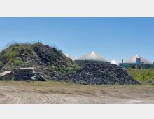 Eine Szene mit mehreren Schutthaufen aus Steinen und Beton im Vordergrund, dahinter drei große, kuppelförmige Gebäude mit weißer Abdeckung, vermutlich Teil einer Industrie- oder Energieanlage, umgeben von Gras und Vegetation. Der Himmel ist klar und blau.