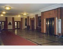 Ein geräumiger Eingangsbereich mit glänzendem Marmorboden, Wänden in warmen Holztönen und großen Türen. An den Wänden hängen Gemälde, und eine rote Läuferbahn verläuft entlang des mittleren Bereichs.