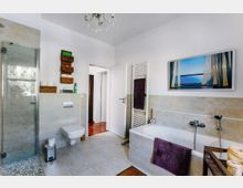 Modernes Badezimmer mit Dusche, Badewanne und WC. Die Wände sind teilweise mit hellen Fliesen verkleidet, es gibt einen Handtuchheizkörper, violette Handtücher und Dekorationselemente wie geflochtene Körbe und ein Bild über der Badewanne.