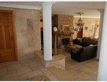 Wohnzimmer in einem rustikalen Stil mit einer gemauerten Kaminwand, einem Kronleuchter, Ledersofas und einem Holztisch. Die Wände sind in einem warmen Beigeton gehalten, der Boden besteht aus hellen Fliesen, und im Vordergrund stehen weiße Säulen.