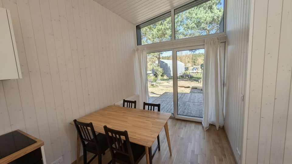 Heller Essbereich in einem modernen Holzhaus mit weiß vertäfelten Wänden, Holzboden und Esstisch mit vier Stühlen. Große bodentiefe Glas-Schiebetür mit Oberlicht und weißen Vorhängen führt auf eine Holzterrasse mit Blick ins Grüne.