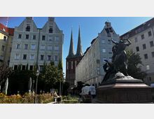 Eine Straßenszene mit einem Denkmal im Vordergrund, das eine Reiterstatue zeigt, flankiert von grünen Gebäuden mit Giebeldächern. Im Hintergrund sind die zwei spitzen Türme der St.-Nikolai-Kirche zu sehen, umgeben von Bäumen, Blumenbeeten und einigen geparkten Fahrzeugen.