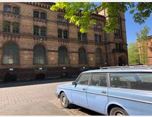 Backsteingebäude mit großen, bogenförmigen Fenstern und dekorativen Zierelementen, umgeben von Kopfsteinpflaster. Im Vordergrund steht ein hellblauer, älterer Kombiwagen, während im Hintergrund Bäume und ein weiteres Backsteingebäude sichtbar sind.