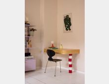 Minimalist home office corner with a light wood floating desk supported by a red-and-white striped column, paired with a black chair on a white plank floor. A lavender shelving unit sits to the left, and a framed graphic artwork hangs on the beige wall above the desk.