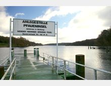 Steg mit Schild „Anlegestelle Pfaueninsel“ über einem ruhigen Gewässer, umgeben von Bäumen an den Ufern. Der Himmel ist teils bewölkt, und im Hintergrund sind weitere Steganlagen sichtbar.