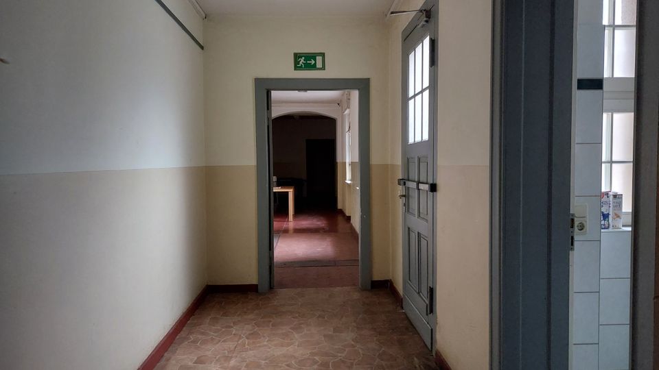 Ein heller Flur mit beige-weißen Wänden und Fliesenboden, der zu einem angrenzenden Raum mit rotem Bodenbelag führt; darüber ein grünes Notausgangsschild. Rechts im Bild eine grau lackierte Tür mit Fenstereinsatz und Blick in einen angrenzenden Raum mit weiß gefliesten Wänden.