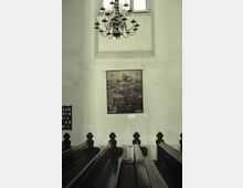 Kircheninnenraum mit Holzbänken, einem großen Kronleuchter an der Decke und einem Gemälde mit religiösem Motiv an der Wand. Im Hintergrund befindet sich ein großes, schlichtes Fenster mit Glasbausteinen.