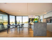 Moderne Innenansicht eines Wohnbereichs mit offener Küche und Essbereich. Große Fensterfronten bieten Blick auf einen Hafen mit Segelbooten, während der Raum mit Holzboden, einer grauen Kücheninsel und Dekorationen wie Pflanzen und Blumen gestaltet ist.