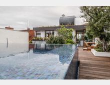 Dachterrasse mit Infinity-Pool, umgeben von Holzboden und Pflanzen. Im Hintergrund ein modernes Gebäude mit großen Glasfronten und einer bepflanzten Dachfläche.