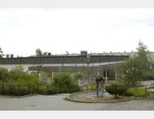 Außenansicht eines flachen Gebäudekomplexes mit grauem Flachdach und verwitterten Fassaden, umgeben von Bäumen und Sträuchern. Im Vordergrund ein gepflasterter Weg, ein Geländer sowie eine kleine Skulptur mit einer Kugel auf Metallstützen.