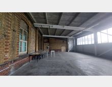 Geräumiger, leerer Innenraum eines Industriegebäudes mit sichtbaren Backsteinwänden, hohen Decken aus Beton und einer Fensterfront auf der rechten Seite, die Tageslicht hereinlässt. Einige Tische und Stühle stehen entlang der Wand auf der linken Seite.