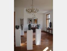 Ausstellungsraum mit Holzboden, in dem drei weiße Sockel mit Kunstskulpturen stehen. An der Wand hängen zwei farbige Bilder, ein Kronleuchter hängt von der Decke, und rechts befindet sich ein Tisch vor einem großen Fenster.