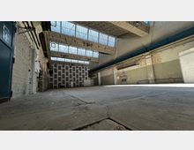 Eine leere Industriehalle mit hohen Decken, freiliegenden Stahlträgern und großen Fenstern, die diffuses Tageslicht hereinlassen. Die Wände bestehen aus weiß gestrichenem Mauerwerk, und links im Vordergrund befindet sich eine blaue Tür.