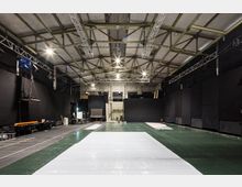 Innenansicht eines großen Proberaums oder Studios mit hohen Decken, sichtbaren Metallträgern und schwarzer Wandverkleidung. Der Raum ist mit Beleuchtungsgerüsten ausgestattet, während auf dem Boden grüne und weiße Abdeckplanen liegen; an den Seiten stehen Tische und Stühle.