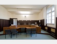 Ein Verwaltungs- oder Sitzungsraum mit Holzvertäfelung an den Wänden, einem halbkreisförmigen Richtertisch mit grauen Stühlen dahinter und mehreren Stühlen davor. Rechts im Raum befinden sich ein Schreibtisch mit Büroausstattung und ein großes Fenster mit weißen Vorhängen.