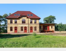 Historisches Bahnhofsgebäude aus gelbem Ziegelstein mit roten Fenstern und Türen. Das zweistöckige Gebäude hat ein rotes Satteldach mit Dachfenstern, eine angrenzende Verladerampe und liegt in einer grünen Umgebung mit Bäumen.